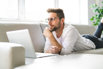 A Young attractive guy is browsing at his laptop, sitting at home on the cozy beige sofa at home, wearing casual outfit