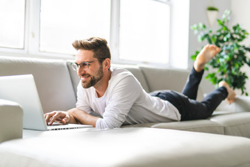 A Young attractive guy is browsing at his laptop, sitting at home on the cozy beige sofa at home, wearing casual outfit