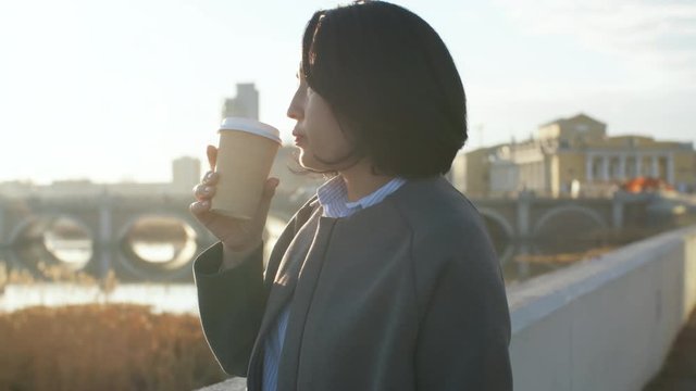 Medium shot of middle-aged Asian woman standing on embankment, enjoying views in the morning and drinking takeaway coffee