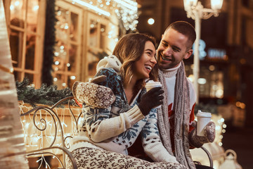 A young romantic couple wearing warm clothes outdoor at Christmas time, sitting on a bench and warming with hot coffee in evening street decorated with beautiful lights.
