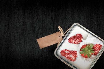 A kitchen food presented on dark black wooden table in a form of a dessert in rectangular glass bowl full of white cream or yoghurt and strawberries with natural paper label.