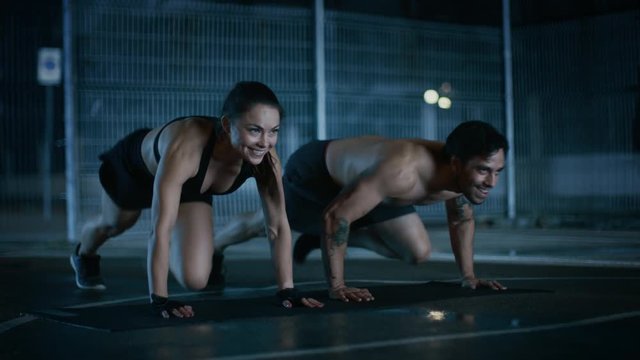 Smiling Happy Athletic Fitness Couple Doing Mountain Climber Exercises. Workout is Done in a Fenced Outdoor Basketball Court. Night Footage After Rain in a Residential Neighborhood Area.