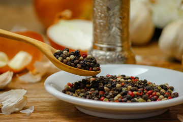 Colored peppers spice mix in spoon and on small ceramic plate. Old wooden table and various of vegetable in the background with red black and white peppercorn.