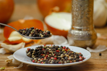 Colored peppers spice mix in spoon and on small ceramic plate. Old wooden table and various of vegetable in the background with red black and white peppercorn.