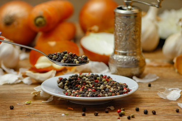 Colored peppers spice mix in spoon and on small ceramic plate. Old wooden table and various of vegetable in the background with red black and white peppercorn.
