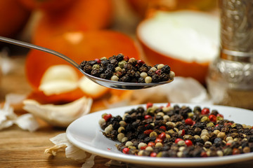 Colored peppers spice mix in spoon and on small ceramic plate. Old wooden table and various of vegetable in the background with red black and white peppercorn.