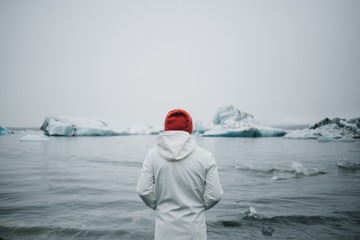 Back of man in white coat or raincoat and red hat, sailor or fisherman stands on beach and looks at...