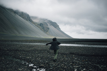 Young man walks on raw and rough cold grey beach with black volcanic sand, wears waterproof jacket for rain protection, take path less travelled, inspiring wanderlust outdoor lifestyle