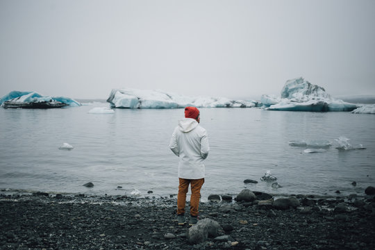 Young man in orange pants, white raincoat and red beanie, tourist stands on black volcanic beach and looks at melting glaciers, climate change danger and realization of responsibility for nature