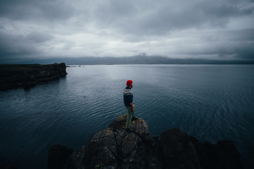 Young bearded hipster man in red fisherman beanie and blue woolen traditional sweater, sailor or explorer stands on edge of rock or cliff, looks at sea, ocean or horizon on cloudy, rainy gloomy day