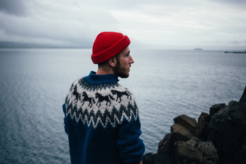 Young handsome man with beard wears traditional authentic blue wool knitted sweater with ornaments and red fisherman or sailor beanie hat, looks to horizon, pensative and thoughtful - obrazy, fototapety, plakaty
