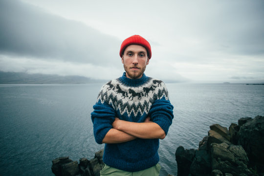 Portrait of serious and tough young bearded man in traditional authentic blue wool sweater with ornaments and red sailor or fisherman beanie stand with crossed arms and look at camera at sealine.