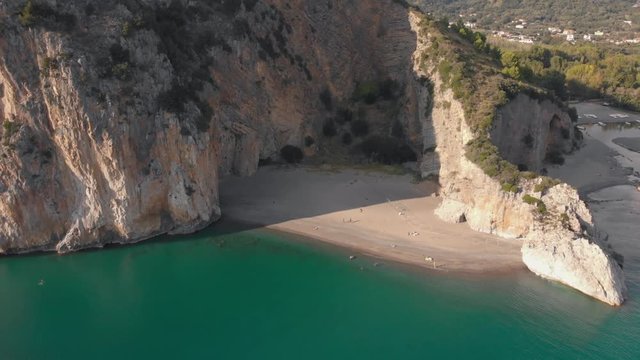 Sandy beach between steep rocks. Mediterranean coastline in sunny day. Aerial, camera moves away