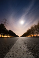Fototapeta na wymiar Empty night asphalt road, bright full moon behind clouds and city light behind two rows of trees on both side of road