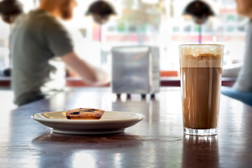 A coffee cappuccino on a transparent glass with a small cake on a wooden table, indoors.
