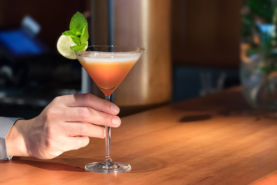 A bartender hand serving a Martini drink on a glass on a wooden counter.