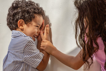 A curly hair little boy looks at his sister laughing as she grabs his arm while he is trying to hide his face.