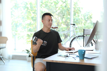 Young man lifting weights and using computer in office. Workplace fitness