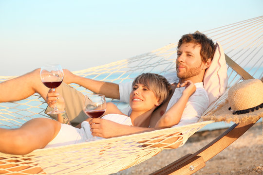 Young Couple Resting With Glasses Of Wine In Hammock On Beach