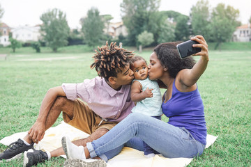 Happy african family taking a selfie with mobile smart phone camera in a public park outdoor - Mother and father having fun with their daughter during a weekend sunny day - Love and happiness concept