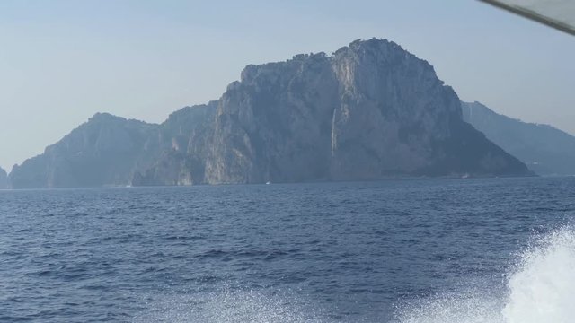 Steep cliff on shores of Capri. Mountain over blue sea. Boat comes up to island. Monte Tiberio, Italy
