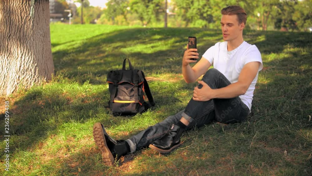 Aufkleber young man sitting in park has video call. caucasian guy using app on smartphone