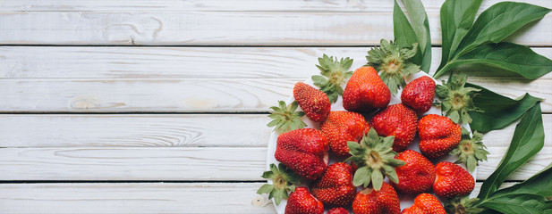 Colorful strawberries on a white ceramic plate with tails and peonies leaves, red berries on a white wooden background. Fresh strawberries on a rustic boards, vegetarian food, copy space, top view.