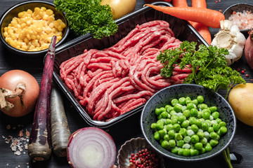 Raw minced meat in container with ingredients for shepherds pie with green peas, yellow corn, carrot, onion and seasonings, on black background, top view