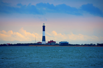 Fire Island Lighthouse with beaming light with Long Island Sound, New York, in the foreground with a colorful sky with dark ominous clouds.