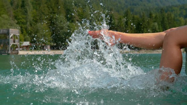 SLOW MOTION CLOSE UP: Unrecognizable playful young woman kicking the beautiful emerald water while enjoying her summer vacation in Slovenian mountains. Carefree girl splashing lake water with her feet