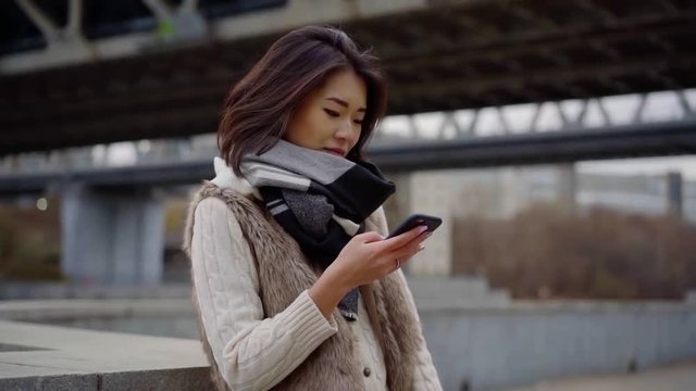 alone chinese woman is waiting someone on city street, surfing internet by smartphone in autumn day
