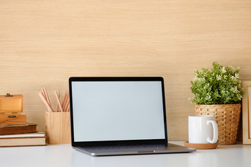 Close-up mockup laptop on white table. books, coffee, pencil. In the background a wood wall and house plants. Empty workplace.