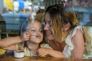 Young mother and daughter eat ice cream in a cafe, happy family, view through the window