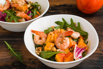 Light fruit salad with fried prawns, persimmon, arugula and walnuts in white bowls on wooden rustic table