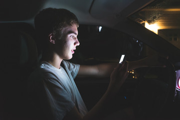 Teenager sitting in his parked car while using his smartphone. He is typing up a message before he begins driving.