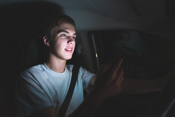 Distracted teenager driving a car with his cell phone in his hand. The light from the screen of the phone is illuminating his face.