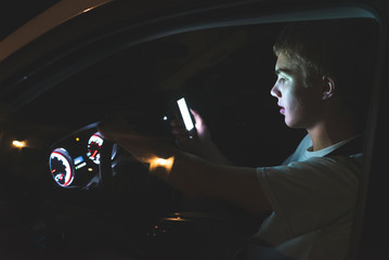 Distracted teenager driving a car with his cell phone in his hand. The light from the screen of the phone is illuminating his face.