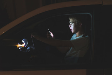 Distracted teenager driving a car with his cell phone in his hand. The light from the screen of the phone is illuminating his face.