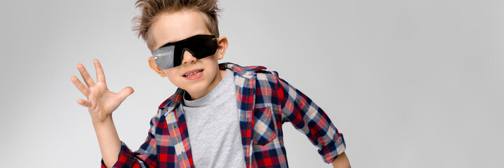 A handsome boy in a plaid shirt, gray shirt and jeans stands on a gray background. The boy in black sunglasses. The boy spread his hands to the sides.