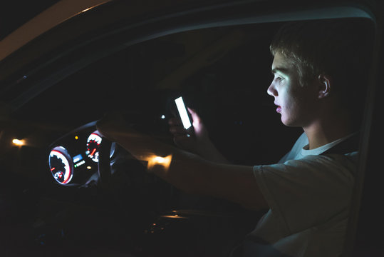 Distracted Teenager Driving A Car With His Cell Phone In His Hand. The Light From The Screen Of The Phone Is Illuminating His Face.