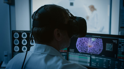 Futuristic Concept: In Medical Control Room Doctor Wearing Virtual Reality Headset Monitors Patient Undergoing MRI or CT Scan Procedure. Computer Displays Show 3D Brain Model with Possible Tumor.