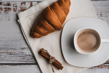 Cup of coffee and a croissant with cinnamon sticks on a napkin on a white wooden table. Morning dinner. Flat lay, view top