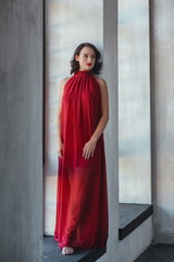 portrait of beautiful young brunette muse woman in red long dress near the window