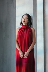 portrait of beautiful young brunette muse woman in red long dress near the window