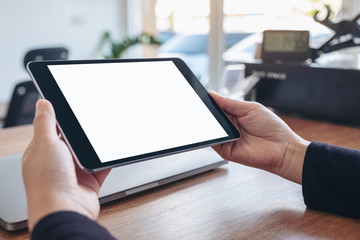 Mockup image of hands holding and using black tablet pc with blank white desktop screen with notebook on wooden table in office