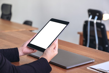 Mockup image of hands holding and using black tablet pc with blank white desktop screen with notebook on wooden table in office