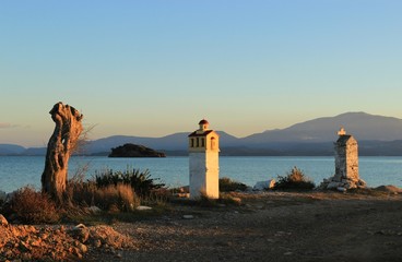 Old dead olive tree two christian orthodox memorial chapels at shore of Ambracian gulf at sunset clear sky view of the blue sea an island and mountains at the horizon at Koronisia village in Greece