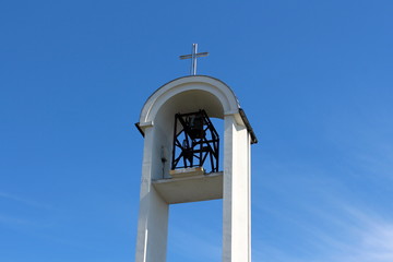 Modern white church bell tower with large open metal bell mechanism, rounded roof and shiny steel cross on top with clear blue sky in background