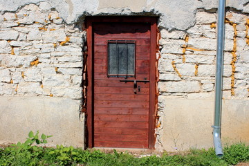 Fototapeta na wymiar New wooden doors made from boards with rusted iron latch and window protection bars mounted on dilapidated cracked stone wall next to new water gutter with uncut grass in front