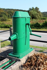 Fototapeta na wymiar Restored old green outdoor metal water pump with large rotating handle mounted on concrete stand surrounded strong metal fence and dried autumn leaves next to paved road with trees and clear blue sky 